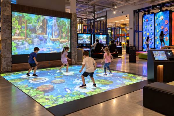 Children playing on an interactive projection floor with digital game screens and interactive attractions in an indoor entertainment center.